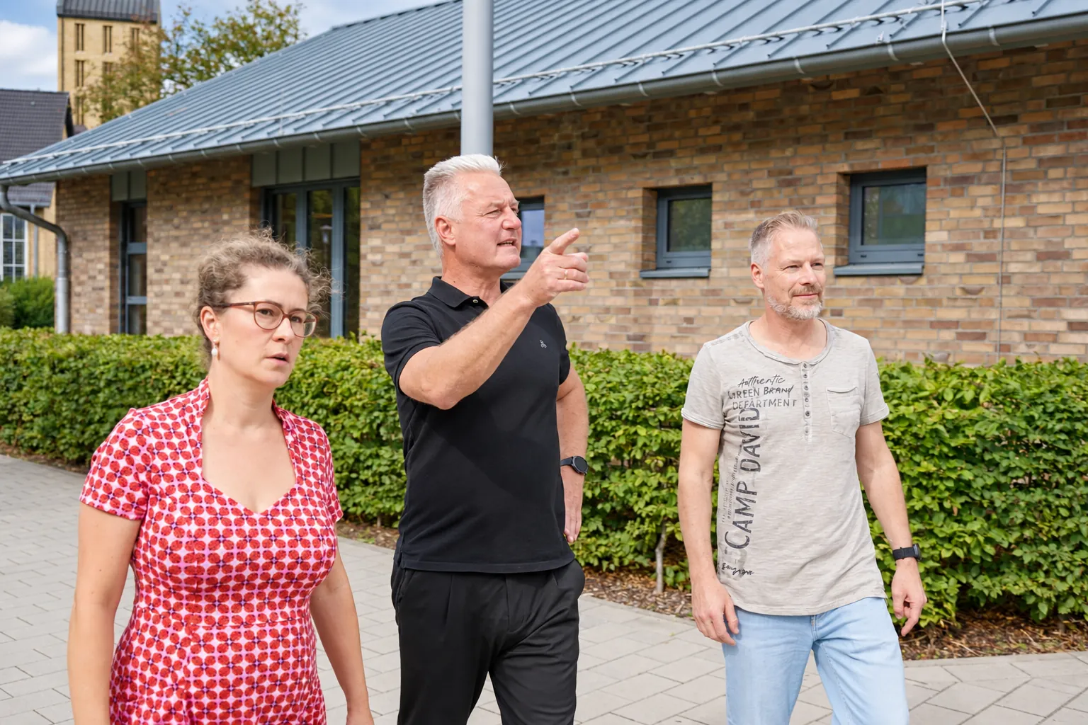 Frank geht mit zwei Menschen einen Weg entlang und zeigt nach vorn.
