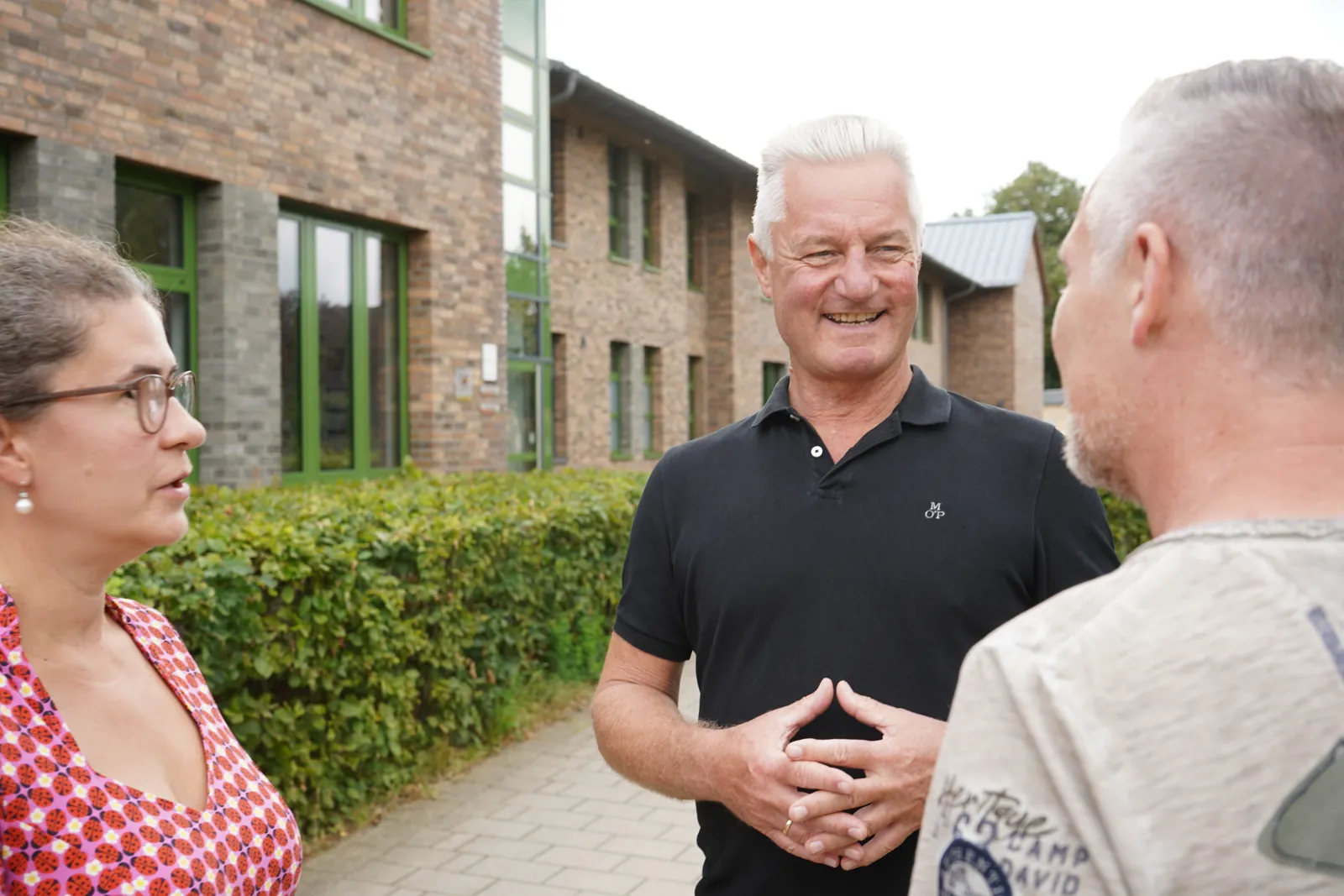 Frank im Gespräch mit zwei Menschen vor einem Backsteingebäude.